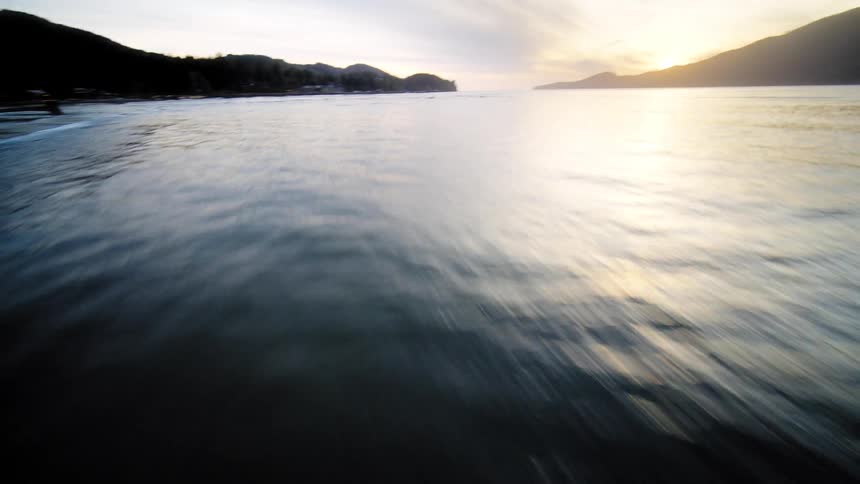 A dynamic FPV drone shot speeds from a sandy beach, across gentle waves, and ascends over a tranquil bay during a beautiful sunrise