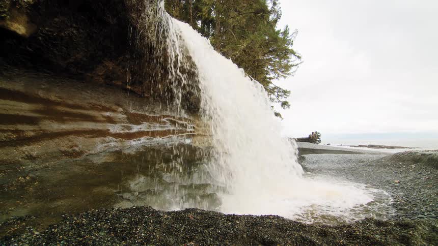 A scenic waterfall cascades from a forest cliff directly onto a rugged, stony coastline