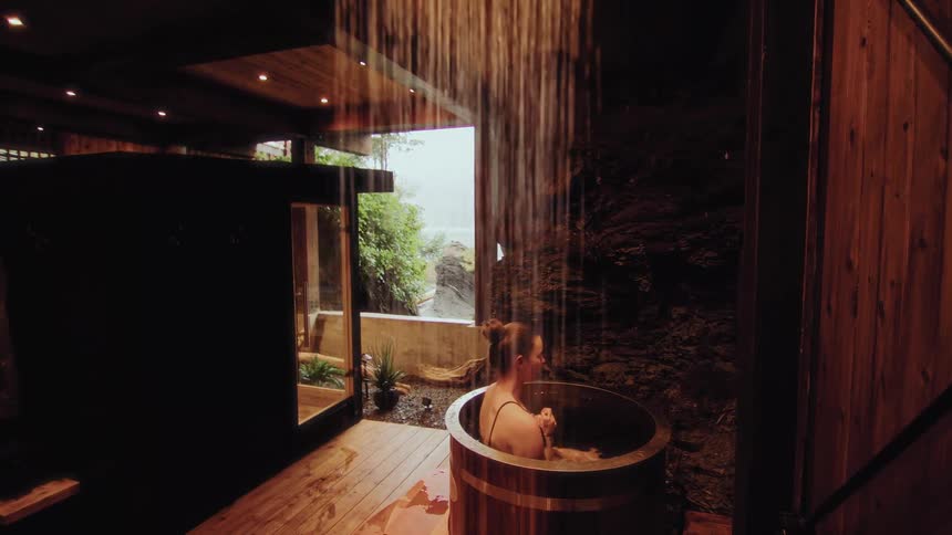 A woman relaxes in a wooden barrel hot tub under a tranquil rainfall shower in a rustic spa with an ocean view