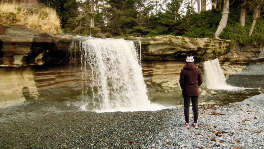 A lone hiker stands in awe before a powerful waterfall cascading over a rocky cliff onto a pebble beach in a remote forest