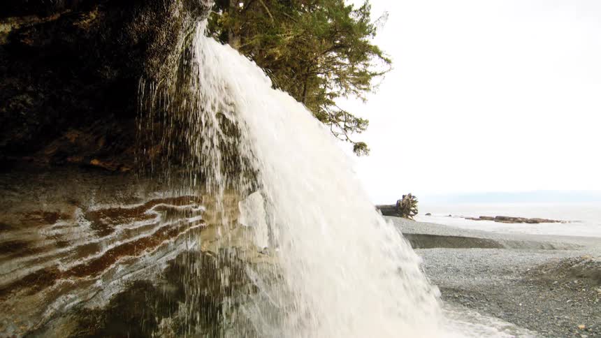 A powerful waterfall cascades over a striped rock formation onto a pebble beach with the ocean in the background