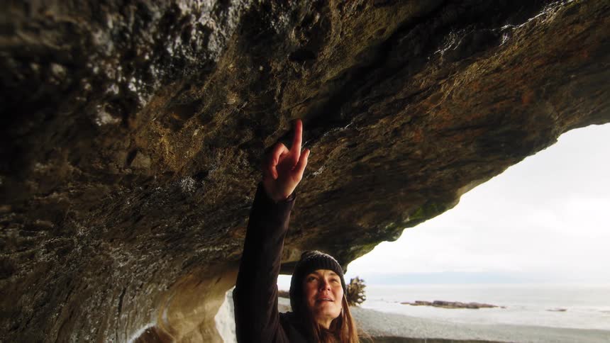 A curious woman in a beanie reaches up to touch the ceiling of a dark sea cave in slow motion