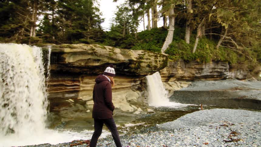 A woman in a winter hat walks in slow motion along a rocky coastline next to a powerful waterfall