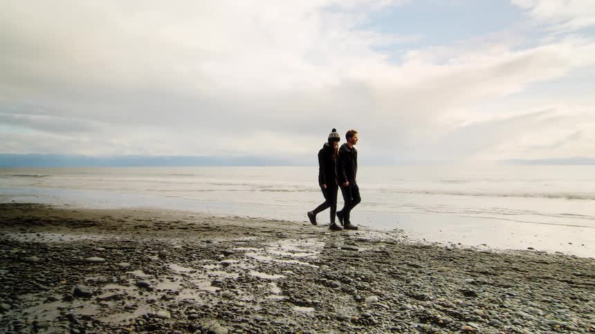 A couple in warm clothes walks along a rocky beach next to the ocean on a cloudy day