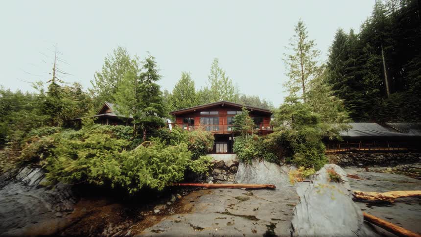 A dramatic reveal shot starts inside a rustic spa with a woman in a swimsuit and pulls back to show the incredible waterfront lodge nestled into the rocky, forested coastline