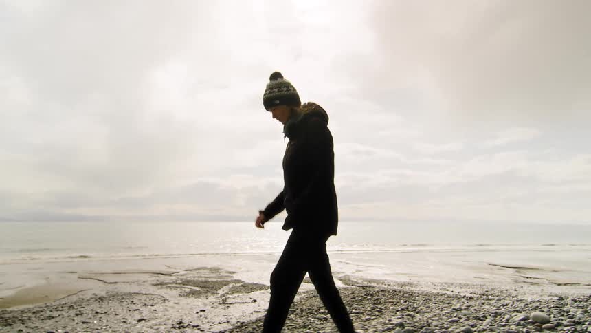 A woman in a winter hat and coat takes a slow, thoughtful walk along a rocky beach on an overcast day