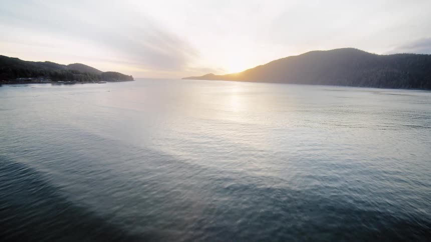 Dynamic aerial shot flying low over a driftwood-covered beach and then soaring out over the calm ocean towards a beautiful sunset between the mountains