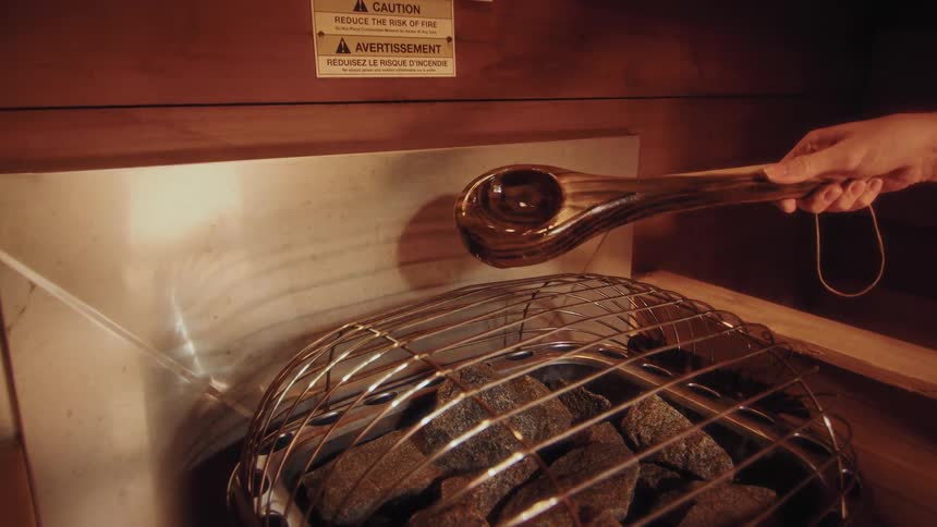 A woman in a traditional wooden sauna pours water from a ladle onto hot rocks to create steam in slow motion