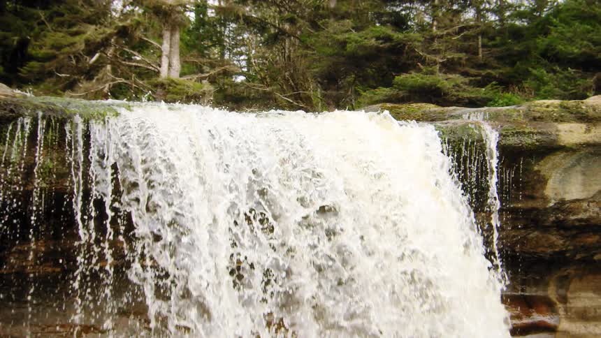 A powerful torrent of water crashes over a wide, rocky ledge in a lush, green forest