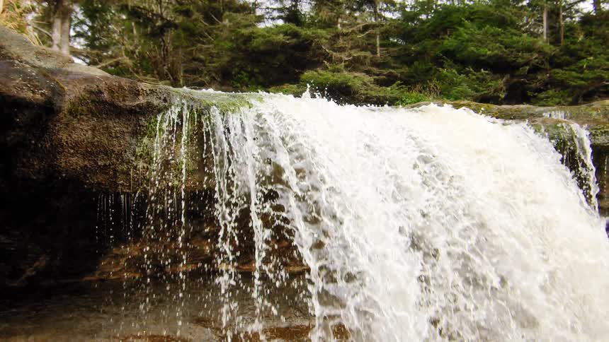 A powerful waterfall cascades over a mossy rock ledge in a lush forest in slow motion