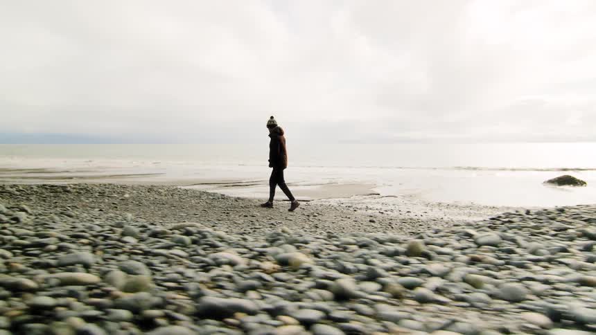A solitary woman wearing a beanie takes a pensive walk along a rocky shoreline on a cold and overcast day