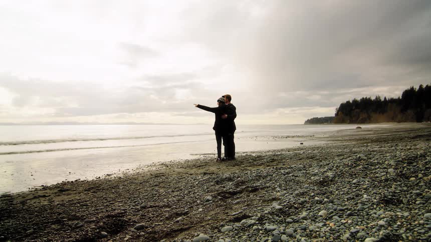 A couple embraces on a rocky shoreline as they watch the ocean waves on a cloudy day