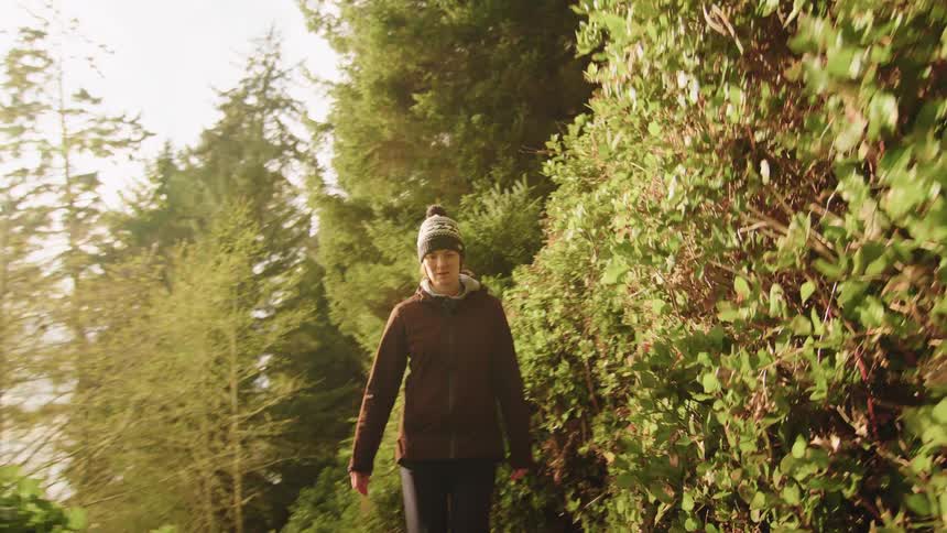 A woman in a beanie slowly walks up a rustic staircase on a sun-drenched forest trail