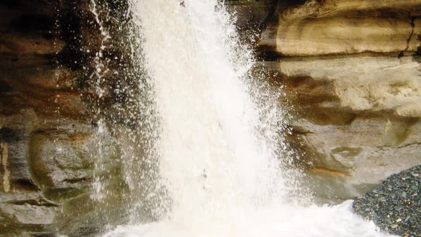Slow motion tilt down a powerful waterfall cascading over mossy rocks and splashing into a tranquil pebble pool