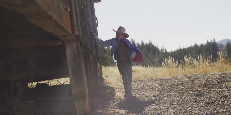 A young boy wearing a cowboy hat and cape plays alongside an old rustic building in a sunny field in slow motion