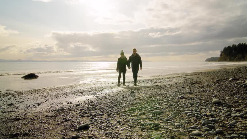 A young couple walks hand-in-hand toward the camera along a beautiful, rocky shoreline on a cool, sunlit day