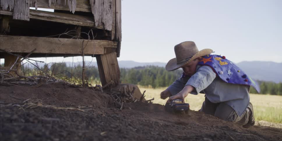 A young boy in a cowboy hat and superhero cape plays with his toy truck in the dirt next to a rustic old shed