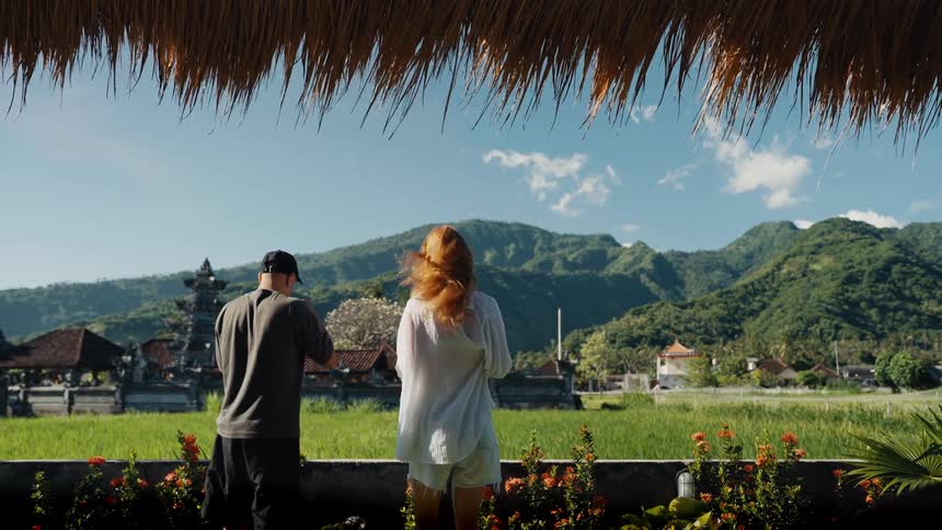 A couple stands under a thatched roof admiring the beautiful view of rice paddies and mountains in Bali