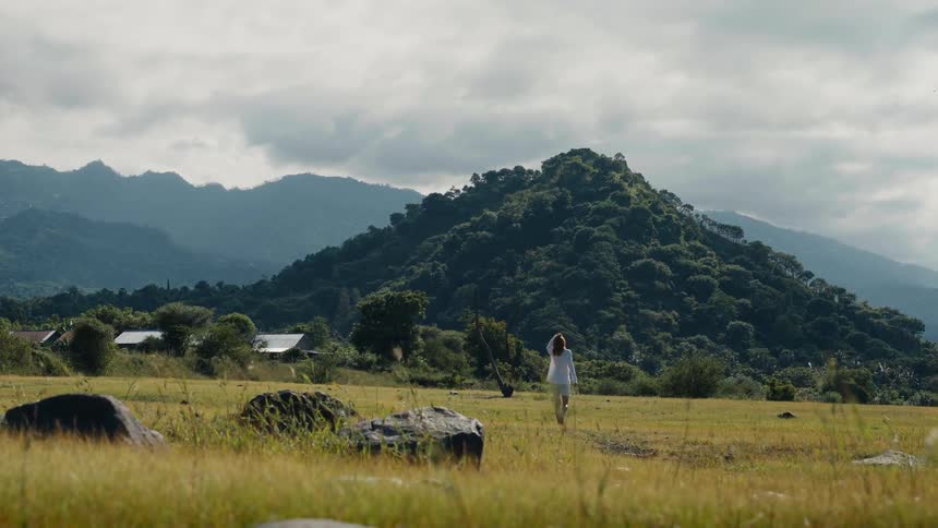 A woman walks through a vast, grassy field, stopping to admire the view of a lush green mountain on a cloudy day