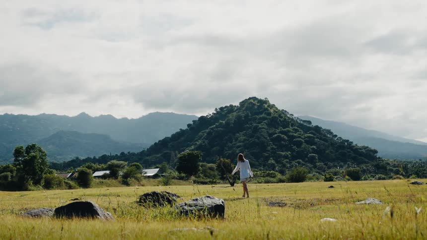 A woman in white strolls through a vast, grassy meadow with a lush, green mountain in the background on a cloudy day