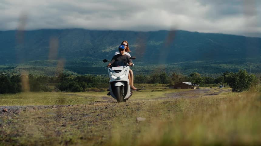 A happy young couple rides a scooter in slow motion along a rural road with stunning green mountains in the distance