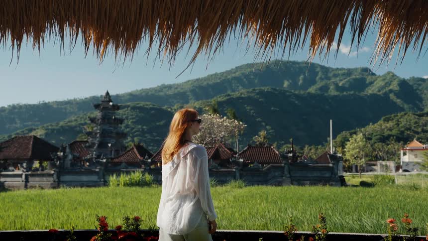 A stylish woman in white enjoys a serene view of a Balinese temple and lush green rice paddies against a mountain backdrop in slow motion