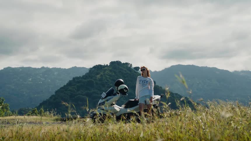 A female traveler stands next to her scooter in a windy field, enjoying the beautiful view of the lush green mountains