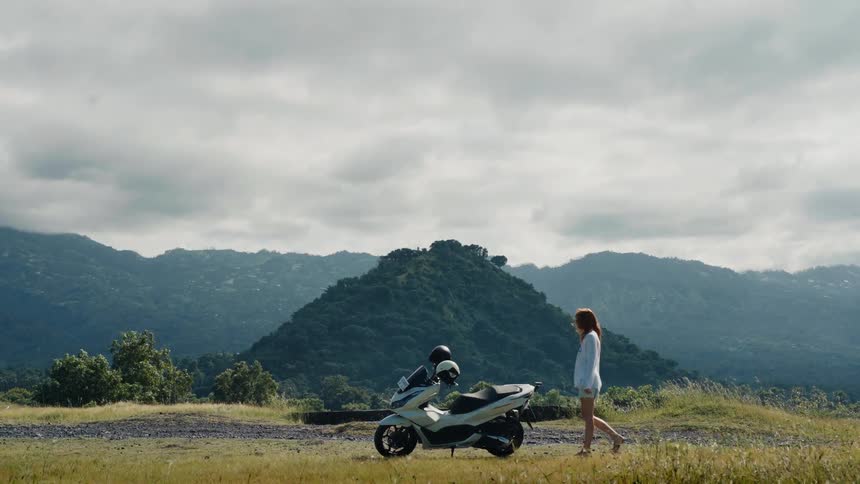A young woman on a road trip walks towards her parked scooter on a scenic hilltop overlooking a lush mountain landscape