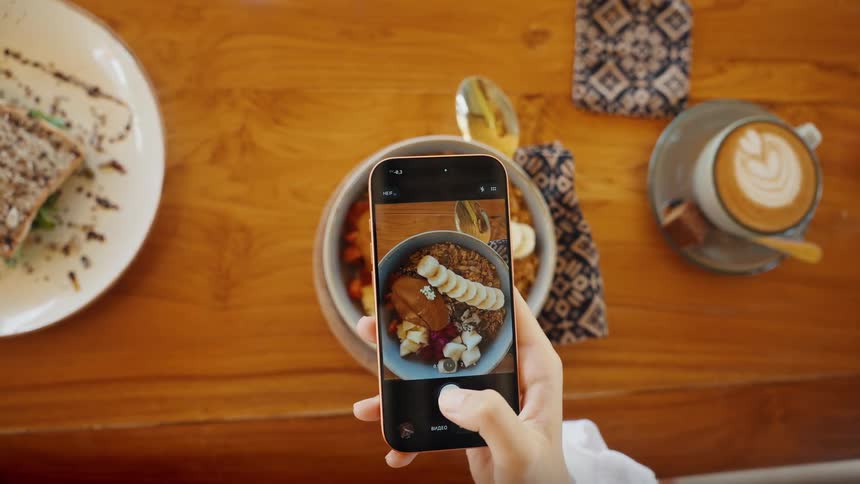 Top-down view of a person using a smartphone to photograph a healthy granola and fruit bowl for breakfast at a cafe