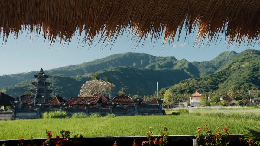 Idyllic view from under a thatched roof of a Balinese temple and lush green rice fields with mountains in the distance