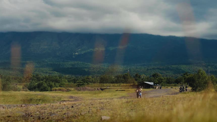 A group of motorcyclists ride on a dirt road through a vast, rural valley with a large green mountain in the background