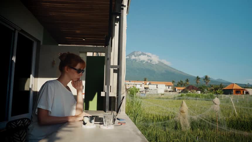 A woman works remotely on her laptop from a sunny balcony with a breathtaking view of a volcano and lush green rice fields