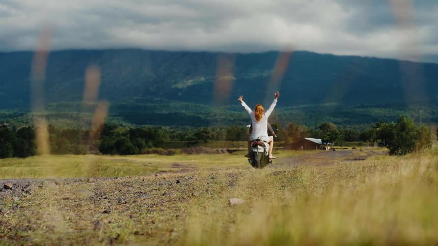 A couple on a scooter rides away down a dirt road towards a vast mountain landscape, the woman spreading her arms in joyful freedom