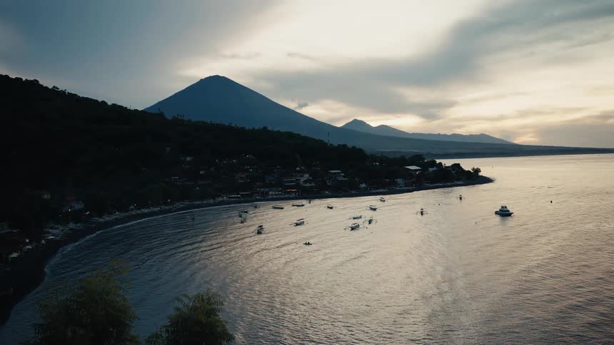 A dramatic sunset over a tranquil bay with fishing boats and a massive volcano towering over the coastline