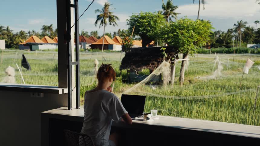 A woman works on her laptop at a bar with a beautiful view of a tropical rice paddy field