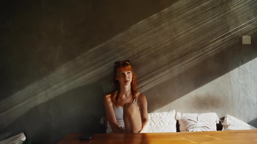A pensive red-haired woman sits alone on a couch, bathed in dramatic diagonal light and shadows from a window