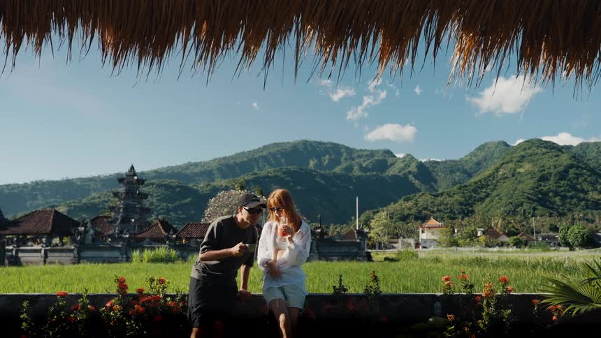 A couple on vacation enjoys drinks together with a stunning view of a Balinese temple, rice fields, and lush mountains