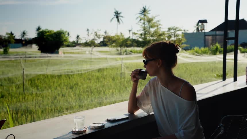 A relaxed woman in sunglasses enjoys a peaceful moment, slowly sipping coffee while gazing at a lush green rice field from a cafe balcony