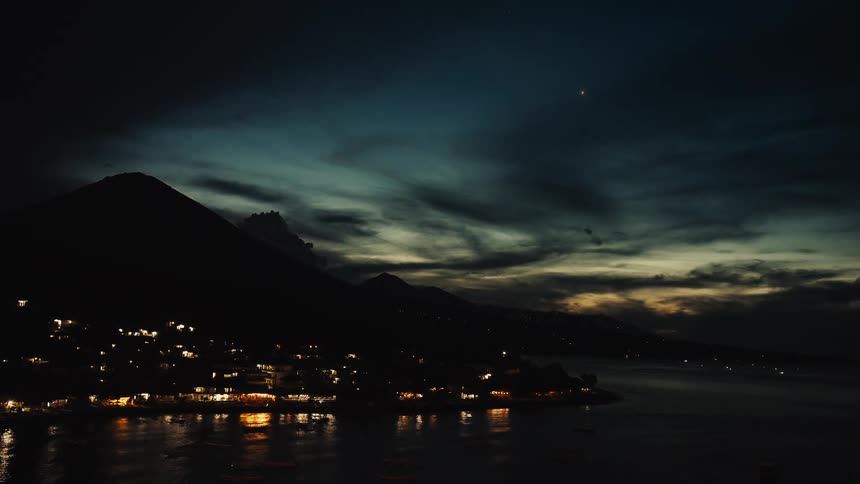 A tranquil view of a lakeside town at night, its lights reflecting on the water below a silhouetted volcano at dusk