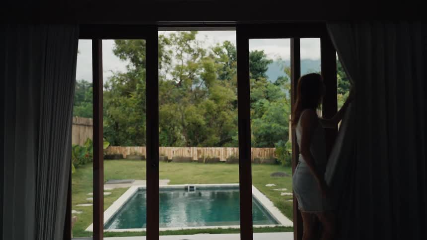 A woman's silhouette is framed as she slowly opens the curtains from a dark room to reveal a lush tropical yard with a private swimming pool