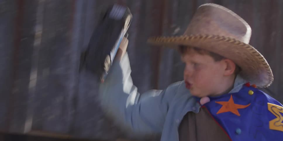 A young boy in a cowboy hat and superhero cape runs in slow motion, playing imaginatively with a toy truck against a wooden wall