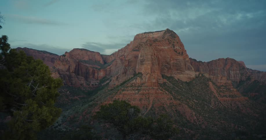 A majestic red rock mountain looms under a cloudy sky at dusk in a vast canyon