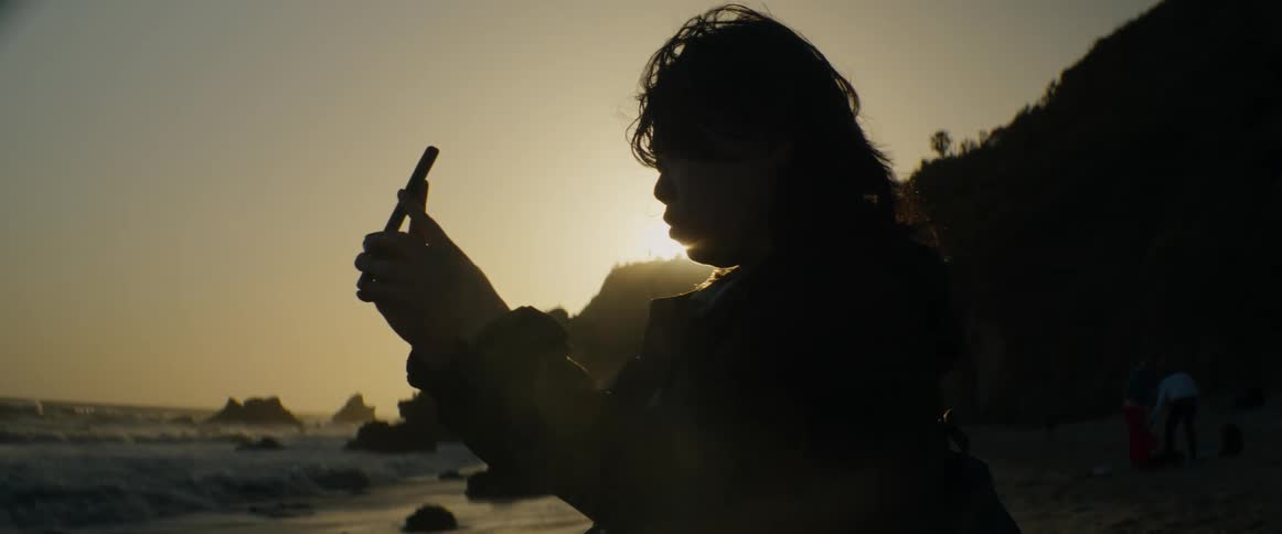 Close up of a person's silhouette as they record the beautiful sunset on a windy beach with their phone