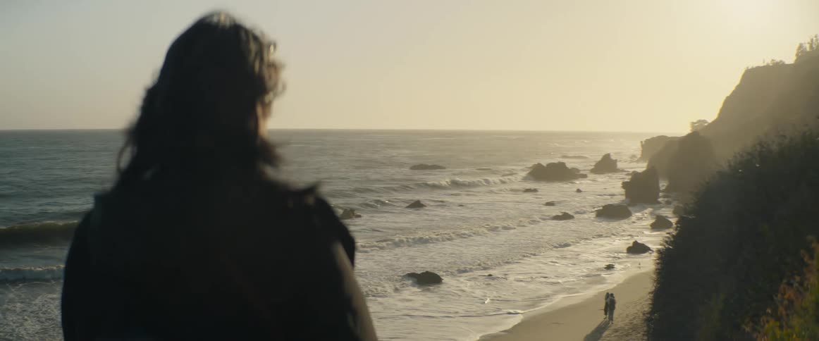 A person with long hair stands on a cliff overlooking a rocky beach with a couple walking on the sand at golden hour