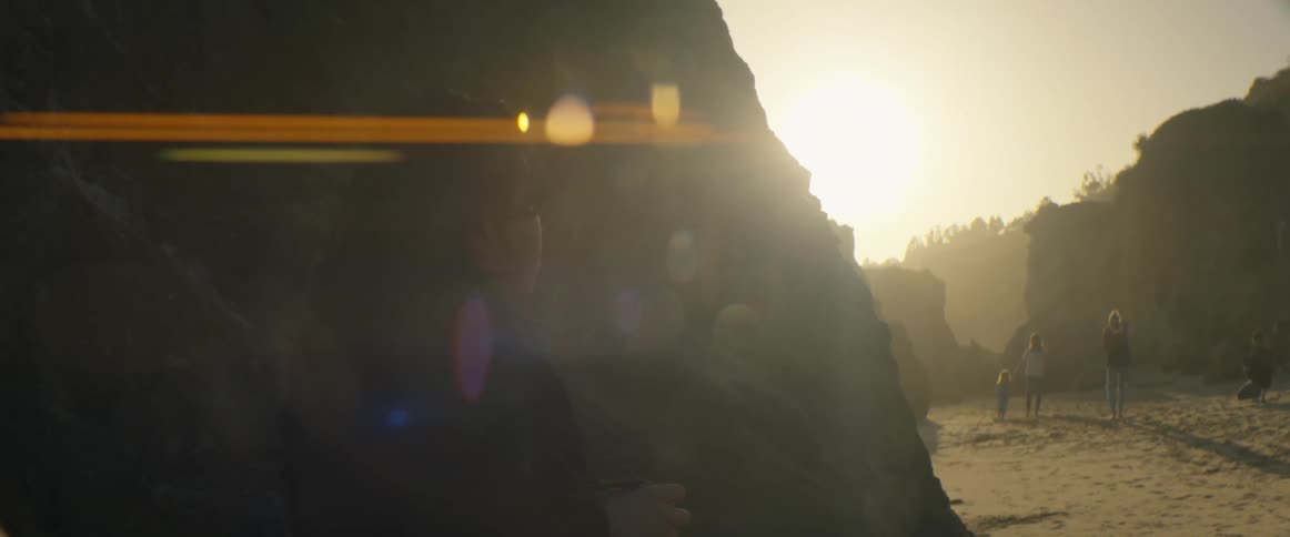 A silhouetted person watches a family walk along a rocky beach in the golden glow of a setting sun with lens flare