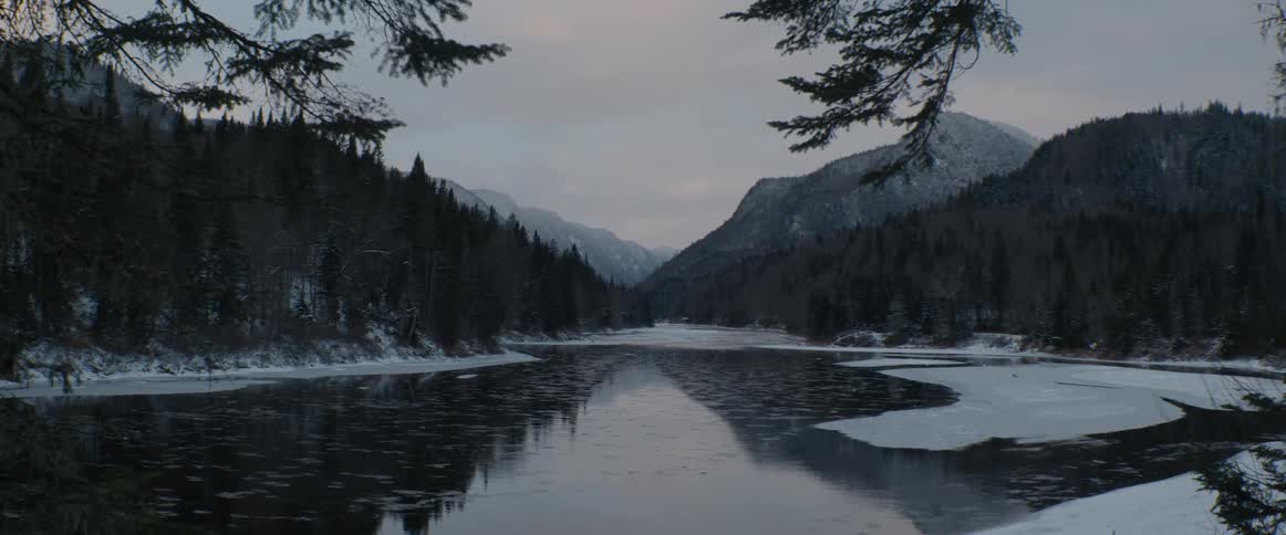A tranquil, partially frozen river winds through a snowy mountain valley during a serene winter dusk