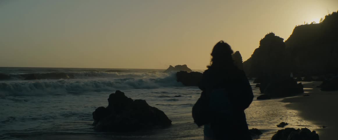 A silhouetted woman stands on a rocky beach at sunset watching the ocean waves crash in slow motion