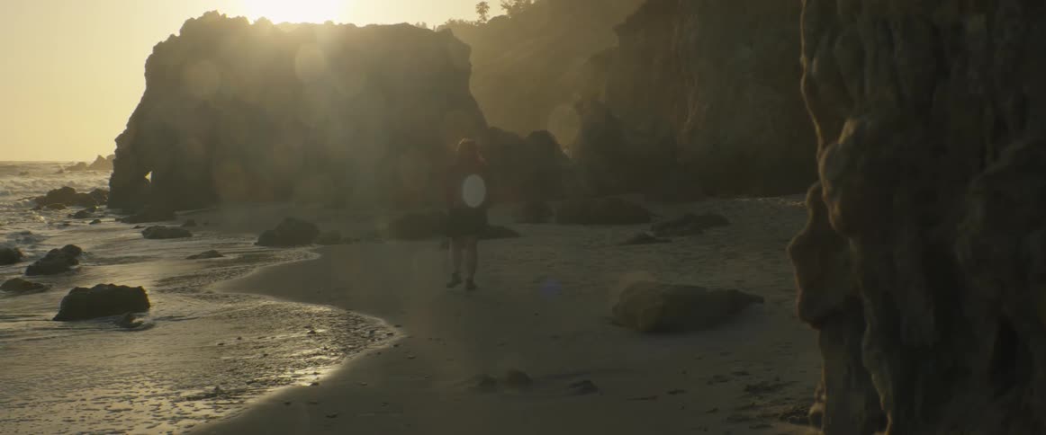 A woman walks alone on a secluded, rocky beach, her silhouette framed by the hazy golden light of the setting sun