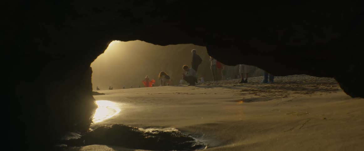 A serene view from inside a dark cave shows people on the beach during a beautiful golden sunset