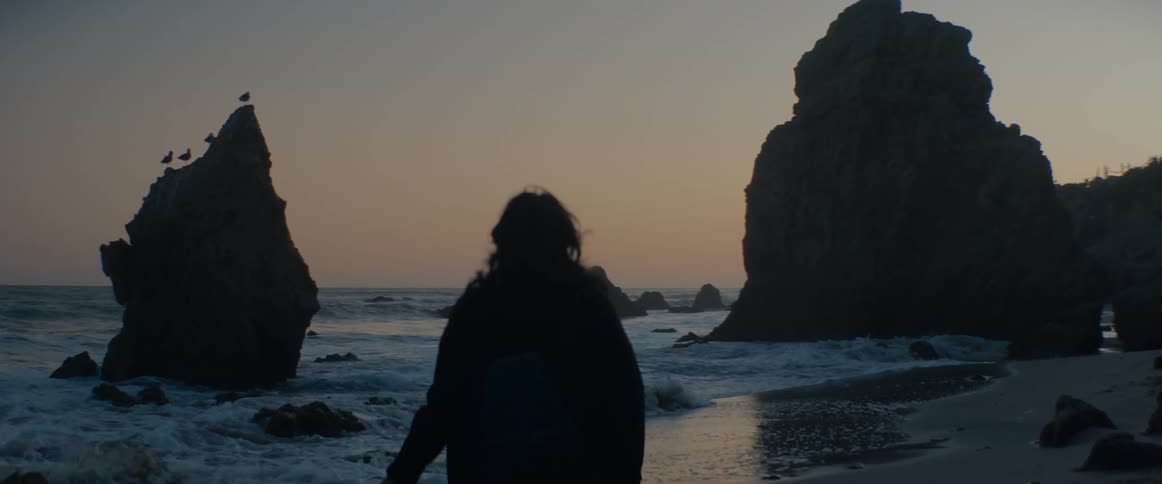Slow motion shot from behind a silhouetted woman as she watches the ocean waves crash on a rocky beach at sunset